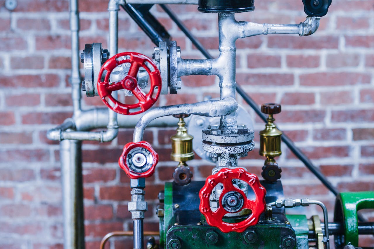 Close-up of industrial machinery with red valve handles and metal pipes against a brick wall.